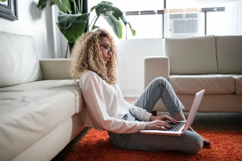 Lady with blonde curly hair and glasses, white blouse and blue jeans sits on floor of lounge with laptop, working from home